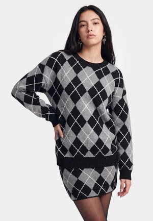 Junge Frau trägt schwarzen und grauen Argyle-Pullover sowie passenden Rock, steht mit einer Hand in der Hüfte vor einfarbigem Hintergrund.