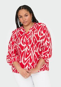 Röd mönstrad blus med vitt abstrakt mönster; har V-ringning och puffärmar, tillverkad av lättviktsmaterial. Matchad med vita byxor.