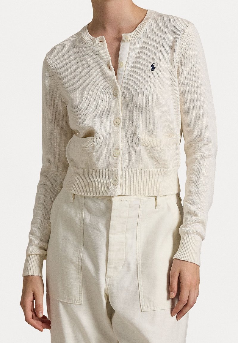 Cardigan color crema con collo rotondo, chiusura con bottoni, polsini a costine e due tasche frontali. Presenta un piccolo logo sul petto.