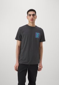 Camiseta de algodón negra con cuello redondo, que presenta un gráfico geométrico en púrpura y verde azulado en el lado izquierdo del pecho. Mangas cortas, corte relajado.