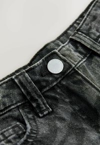 Lähikuva mustista denim-farkkujen vyötäröstä, jossa on metallinappi, vyölenkit ja näkyvät ompeluyksityiskohdat.