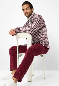 Rode corduroybroek en een bordeauxrode en witte geruite shirt, gecombineerd met witte sneakers, zittend op een witte stoel tegen een lichte achtergrond.