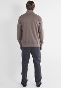 Veste en polaire marron avec col haut, poignets et ourlet côtelés, associée à un pantalon gris foncé. Design simple, sans motifs ni logos visibles.