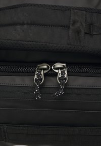 Zwarte rugzak van textuurstof, dubbele ritssluiting met metalen hardware en versterkte webbingdetails. Glad en duurzaam materiaal.