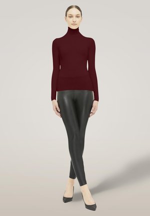 Kvinde står med benene krydset, iført en tætsiddende bordeaux rullekravebluse, sorte læderleggings og sorte høje hæle.