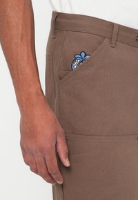 Pantaloni marroni in cotone con un tessuto texture. Presentano una tasca curva con un design ricamato blu. Mano appoggiata di lato.