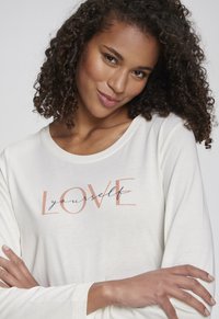 Langarm-Cremeshirt mit Rundhalsausschnitt, auf dem der Text "LIEBE dich selbst" in einer Mischung aus roségoldenen und schwarzen Schriftarten zu sehen ist. Glatte Oberfläche.