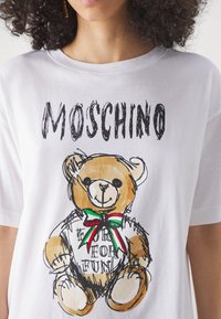 Kobieta ubrana w białą koszulkę z rysunkowym misiem i napisem "MOSCHINO" powyżej, napisanym grubymi literami.
