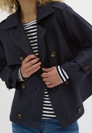 Kvinde, der justerer en marineblå dobbeltradet frakke med brune knapper over en sort- og hvidstribet skjorte og blå jeans mod en ensfarvet baggrund.