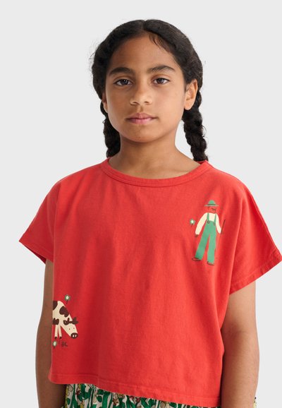 Jeune fille aux cheveux tressés portant un t-shirt rouge avec une illustration d'un fermier tenant une fourche et une vache en noir et blanc.