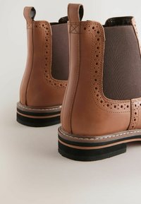 Bruine leren Chelsea-laarzen met brogue-details, elastische zijpanelen, aantreklussen en gestapelde zwarte en bruine zolen op een lichte achtergrond.