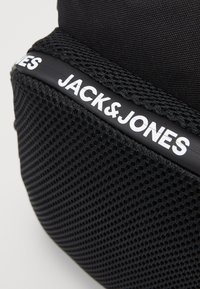 Czarna torba tekstylna z panelami z siatki, z wyraźnym białym logo "JACK&JONES" na błyszczącym czarnym akcentem. Gładka tekstura i wyraźny kształt.