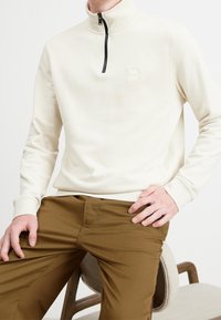 Béžový pulover na půl zip vyrobený z texturované látky, s malou nášivkou loga a žebrovanými manžetami, v kombinaci s hnědými kalhotami.