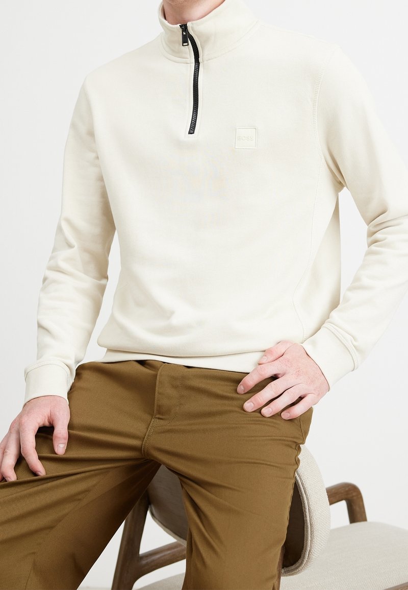 Béžový pulover na půl zip vyrobený z texturované látky, s malou nášivkou loga a žebrovanými manžetami, v kombinaci s hnědými kalhotami.