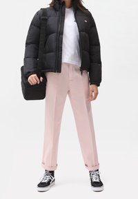 Czarna pikowana kurtka z wysokim kołnierzem, biała koszulka, różowe szerokie spodnie, czarne sneakersy z białymi akcentami; niesie czarną torbę.