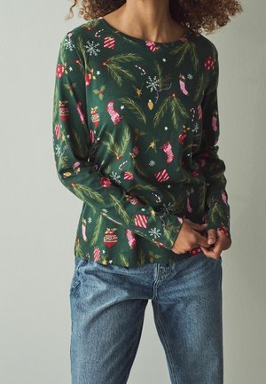 Femme portant un t-shirt à manches longues vert foncé avec des motifs de Noël et un jean bleu, debout, les mains jointes devant elle.