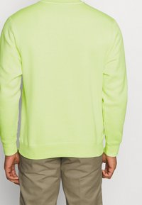 Neongrön sweatshirt med långa ärmar, ribbad mudd och nederkant; slät textur, rund halsringning; bärs med beiga byxor.