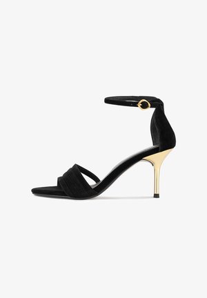 Schwarze Wildleder-Sandalen mit hohen Absätzen in Goldoptik, ausgestattet mit einem Knöchelriemen und gekreuztem Riemen über den Zehen.