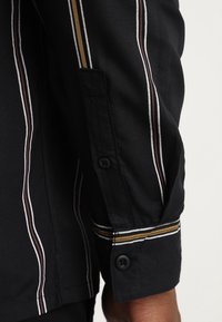 Zwart shirt met verticale strepen in wit, bruin en goud. Gedetailleerde manchet met knoopsluiting. Gladde stoftextuur.
