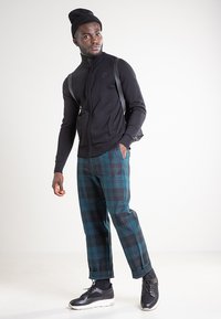 Zwarte jas met rits, groen-zwart geruite broek, zwarte sneakers en een gebreide muts. Heeft een aansluitend ontwerp en gestructureerde materialen.