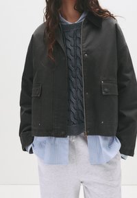 Veste noire courte avec détails de boutons et deux poches avant, portée sur un pull bleu en câble tricoté et une chemise légère rayée. Pantalon de survêtement gris clair.