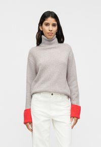 Ανοιχτό γκρι πουλόβερ με ζιβάγκο και ριμπ μανσέτες σε φωτεινό κόκκινο. Μαλακή υφή, oversized εφαρμογή, συνδυασμένο με άσπρο παντελόνι. Απλός και μοντέρνος σχεδιασμός.