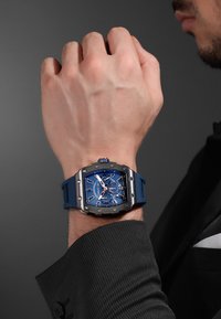 Reloj azul con caja metálica cuadrada, correa de goma y esfera multisanal con acentos plateados y fondo azul. Lleva puesto en una muñeca.