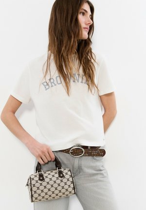 Mujer joven con cabello largo y castaño, que lleva una camiseta blanca con la palabra "BROWNI", vaqueros gris claro, cinturón marrón con tachuelas y sostiene un bolso con estampado.