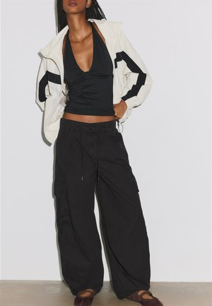 Femme portant un top dos-nu noir, une veste blanche et noire, un pantalon cargo noir ample et des chaussures marron foncé, debout contre un mur blanc.