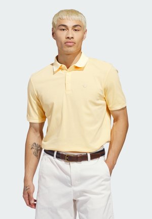 Pikétröja i pastellgult bomull, med krage, korta ärmar, tre knappar och en liten logotyp på bröstet. Kombinerad med vita byxor.