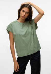 Žalias medvilninis marškinėliai laisvu siluetu, su nėriniuota apdaila pečių siūlėse. Trumpomis rankovėmis ir apvalia iškirpte.