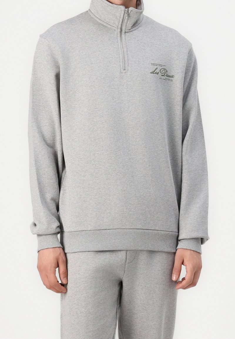 Sweat-shirt gris à demi-fermeture éclair, fabriqué en matériau doux, avec un col et des poignets côtelés, et un détail subtil du logo sur la poitrine.