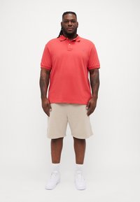 Polo shirt rouge, à manches courtes, avec col, associé à un short beige. Le mannequin porte des baskets blanches. Design simple, texture semblable à du coton.