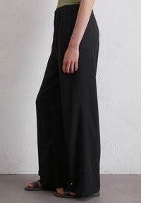 Zwarte wijde broek gemaakt van een gladde stof met een elastische tailleband, met een ontspannen pasvorm en omgeslagen broekspijpen. Draagt met zwarte sandalen.