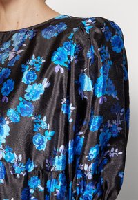 Tissu en satin noir avec un motif floral bleu et violet, présentant une couture froncée et une épaule légèrement bouffante sur un vêtement.