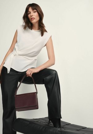 Biała bezrękawnik z detalem w postaci węzła, czarne skórzane spodnie oraz burgundowa skórzana torebka, wyeksponowane na czarnej teksturowanej powierzchni.