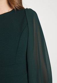 Tumman vihreä mekko, jossa on rakenteellinen yläosa ja pehmeät, läpikuultavat hihat, joilla on sileä tekstuuri. Hihoissa on hienovarainen kerätty yksityiskohta.