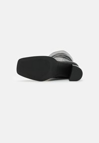 Botte noire en cuir montant jusqu'au genou avec une tige lisse et une semelle en caoutchouc texturée. Dotée d'un bout carré et d'un talon de hauteur moyenne.