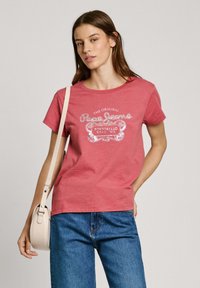 T-shirt en coton rouge à manches courtes, avec impression de texte et de design blancs. Assorti à un jean en denim bleu et à un sac en bandoulière couleur crème.