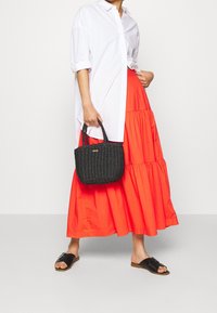 Nainen, jolla on yllään valkoinen napitettu paita, pitkä kirkkaanpunainen kerroksellinen hame, mustat sandaalit ja joka pitää mustaa kudottua käsilaukkua sivullaan.