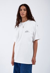 Camiseta de algodón blanca con un corte relajado. Presenta una firma negra en el lado izquierdo del pecho y mangas cortas. Combinada con jeans azules.