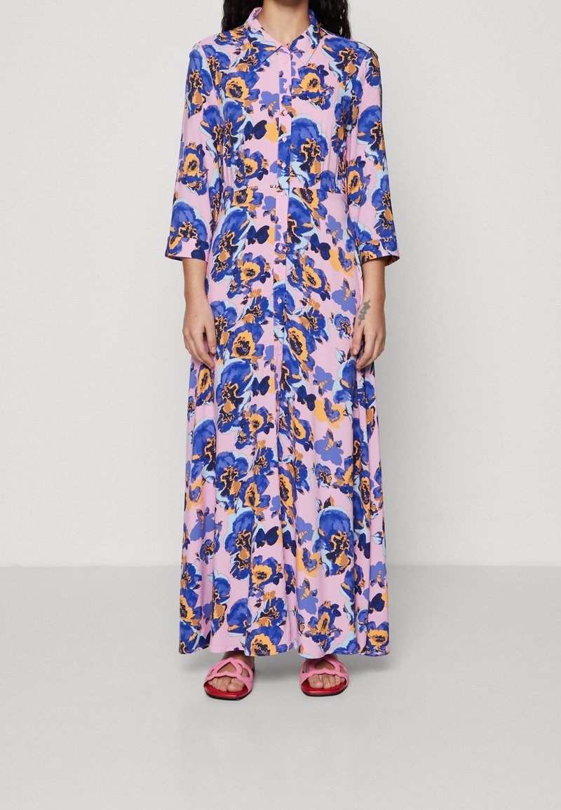 Długa sukienka w kwiaty z różowym tłem oraz niebieskimi i żółtymi wzorami, z zapięciem na guziki z przodu, kołnierzykiem i rękawami o długości trzech ćwiartek, połączona z różowymi sandałami.