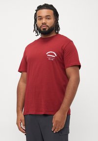 Rød bomulds t-shirt med korte ærmer, der har hvid tekst på venstre bryst og et afslappet pasform design. Enkel, ubesværet stofstruktur.