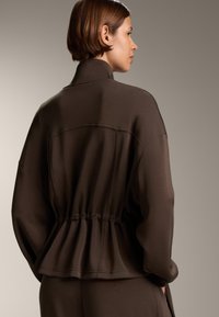 Braune, leichte Jacke mit kragenloser Halslinie, tailliertem Bund und weiten Ärmeln. Glatte Textur mit einem subtilen Glanz. Schlichtes Design.