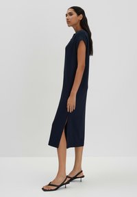 Mittelblaues Midi-Kleid mit kurzen Ärmeln, seitlichen Schlitzen und glattem Stoff. Kombiniert mit schwarzen, römischen Sandalen auf einem neutralen Hintergrund.