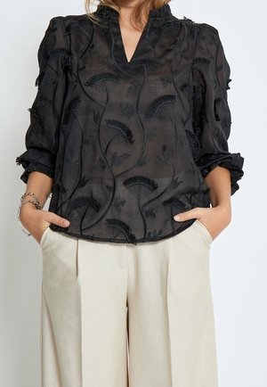 Femme portant un chemisier noir transparent brodé de fleurs avec des manches bouffantes et un pantalon beige clair à jambes larges, les mains dans les poches.