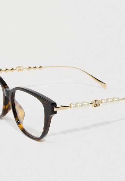 Armação de óculos quadrada em tartaruga com hastes douradas decoradas com cinco pérolas e um pequeno enfeite circular perto das dobradiças.