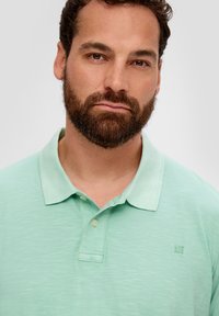 Jasnozielona koszulka polo z krótkim rękawem, o teksturowanej tkaninie, z trzema guzikami i małym haftowanym logo na piersi.