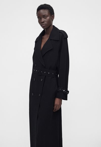 Μαύρη γυναίκα με μακρύ μαύρο διπλόμπροχο παλτό με ζωνάρι και λουράκια στα μανίκια, στέκεται μπροστά σε μονόχρωμο ανοιχτό γκρι φόντο.