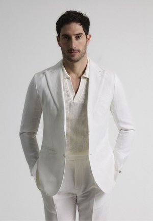 Chaqueta de traje blanca de lino con solapas de pico, llevada sobre un polo de punto texturizado ligero. Corte entallado, combinado con pantalones a juego.